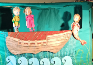 W łodzi stoi chłopiec z dziewczynką. Z prawej strony siedzi syrenka.